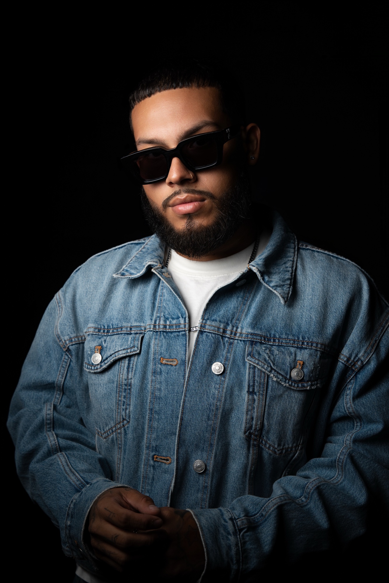 Kae G in a light denim jacket and black sunglasses, hands clasped low, chin slightly down