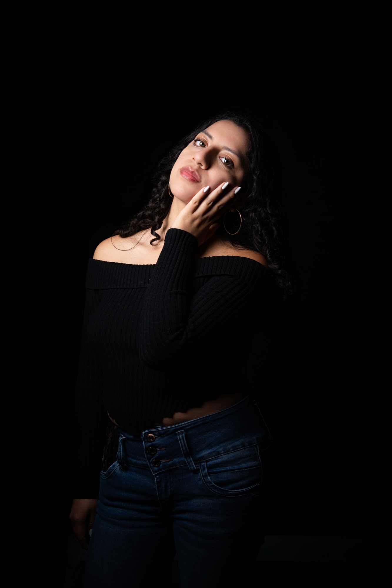 Elé in a black off the shoulder sweater and jeans, one hand raised to her cheek