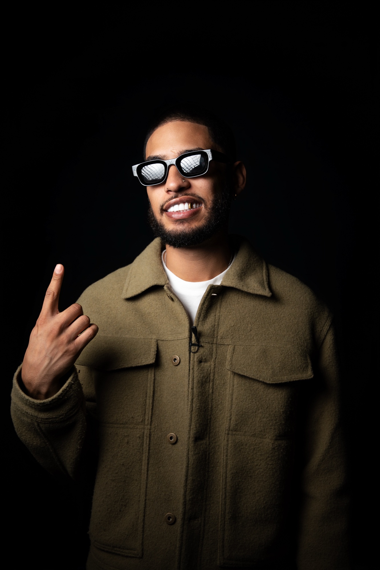 E1even in an olive shirt jacket and pixel sunglasses, one hand raised, smiling at the camera