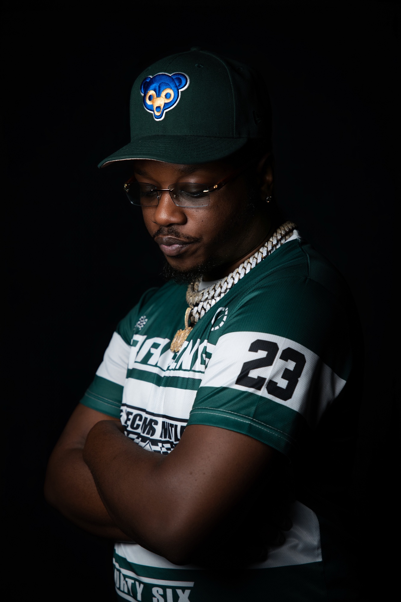 Que Billionaire in a green number 23 jersey and matching cap, arms folded, looking down