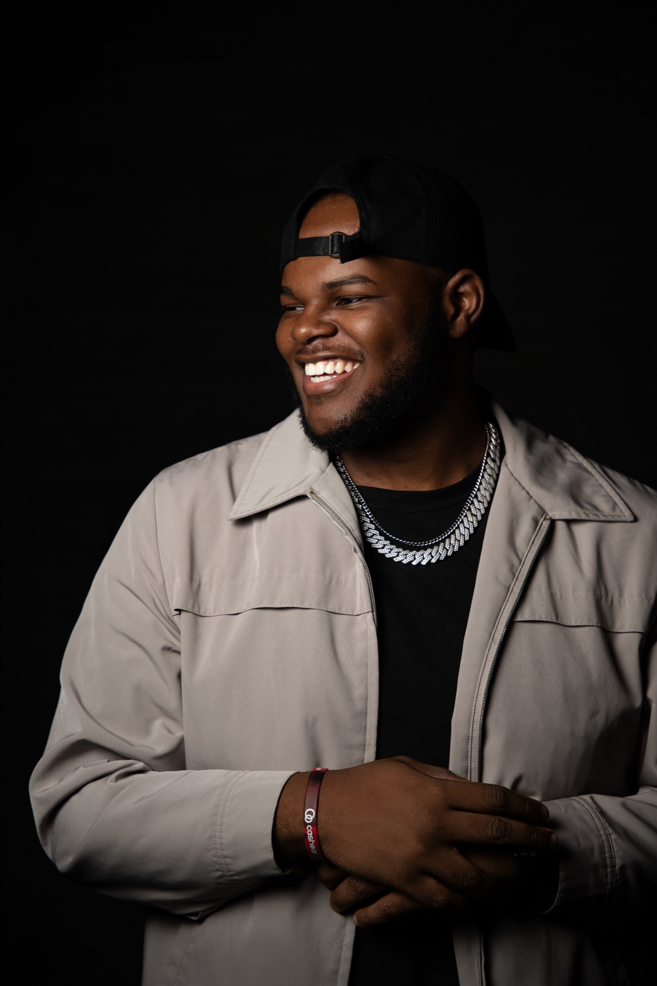 Ali X laughing in a grey jacket and backward cap, silver chain, looking off to the side