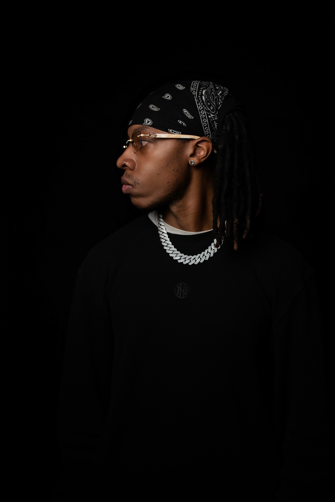 Jayneto in profile wearing a black bandana, glasses and a silver chain over a black crewneck