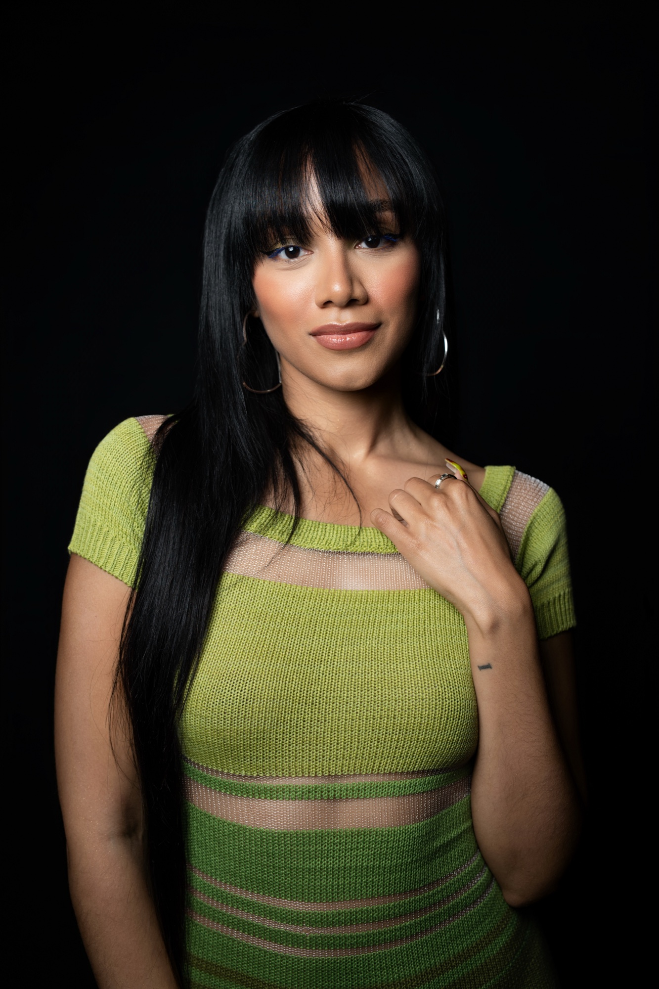 Ame Luv in a green knit top with hoop earrings, one hand resting at her collarbone
