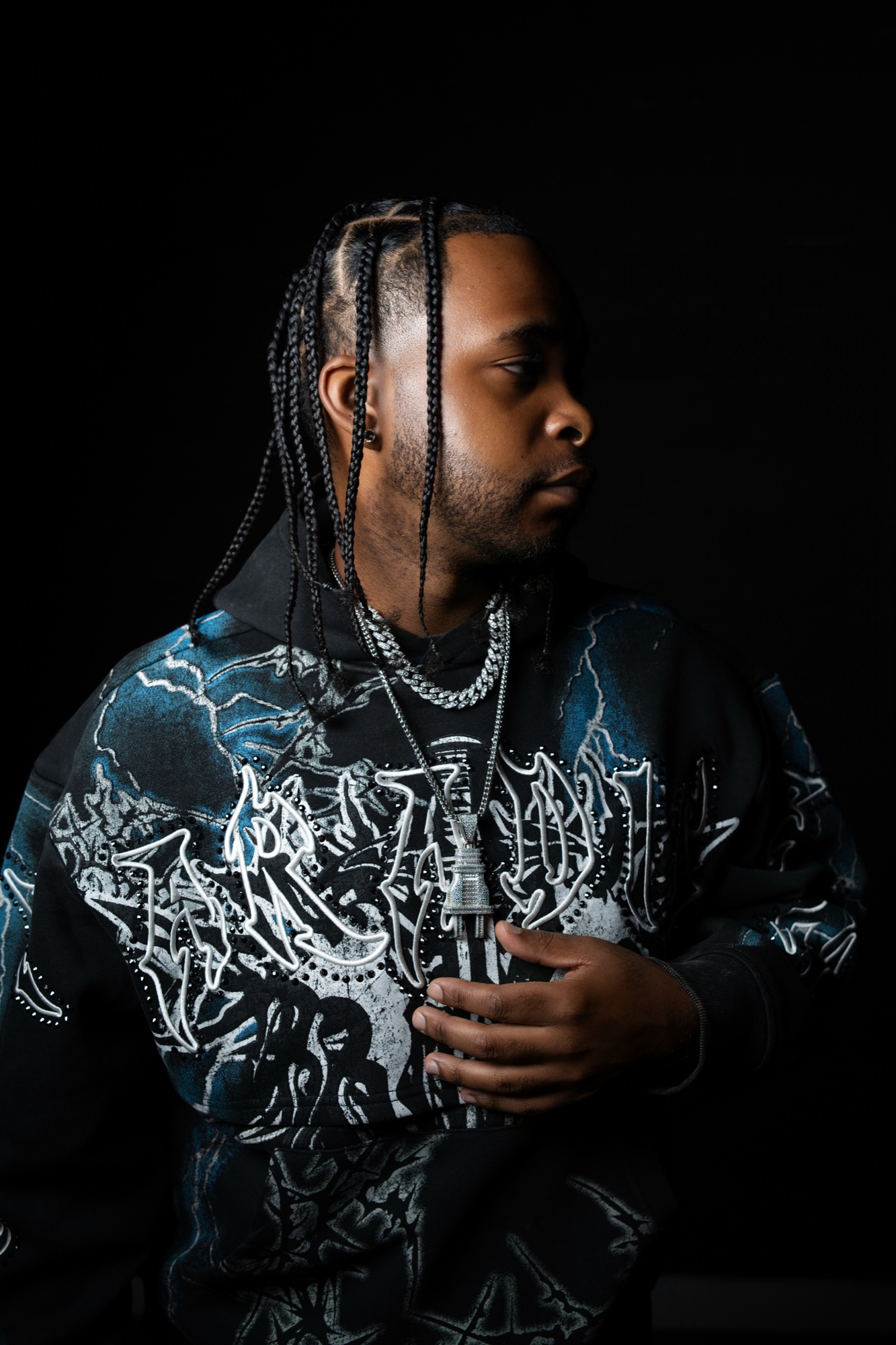 Platnume in a dark graphic hoodie, braids back, holding a pendant and looking to the side