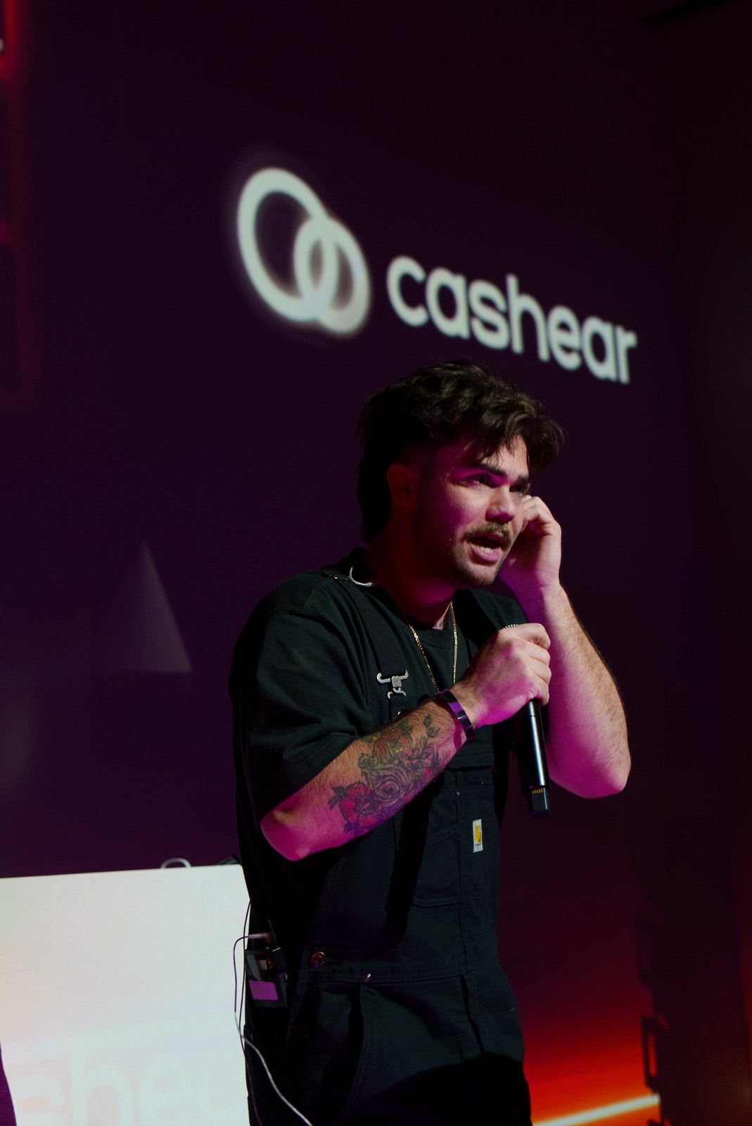 A man in a black shirt with tattooed forearms speaks into a microphone beneath a projected Cashear logo