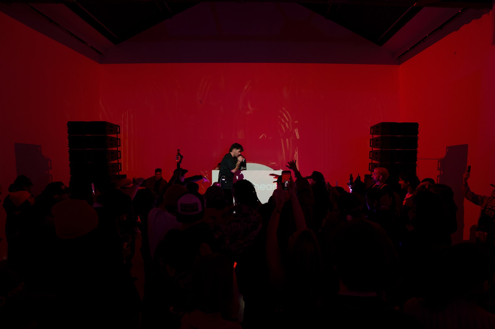 Vocalist performs at a white booth inside a room lit red and lined with tall speaker stacks