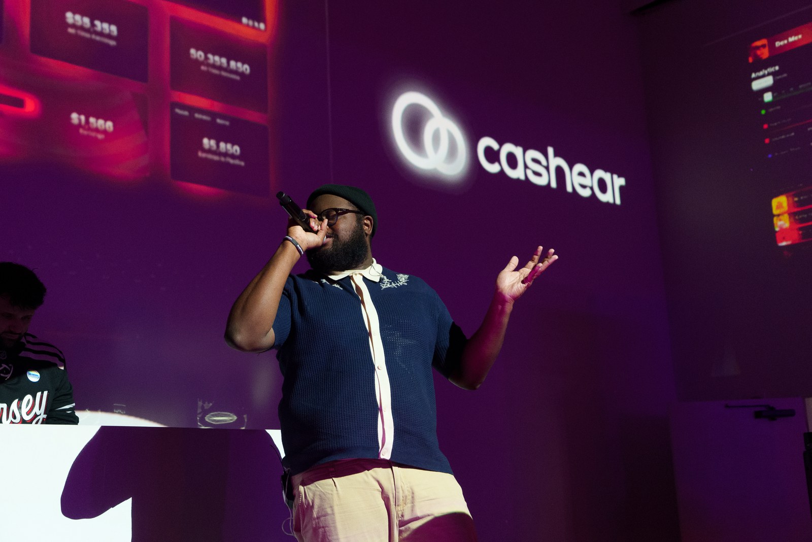 Performer in a navy polo and cap singing into a microphone before a purple Cashear projection