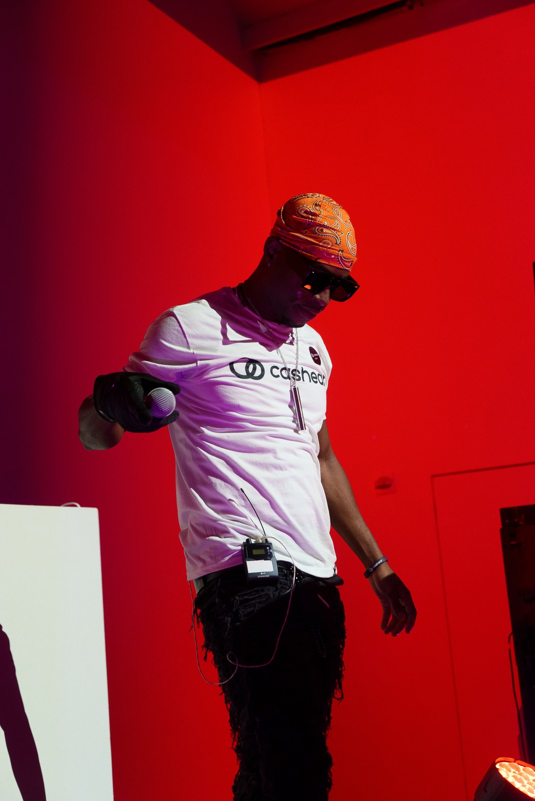 A performer in an orange bandana, sunglasses and a white Cashear shirt holds a microphone against a red wall
