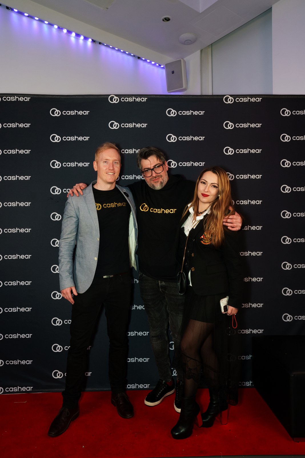 Three guests pose arm in arm on a red carpet in front of a black Cashear step and repeat banner