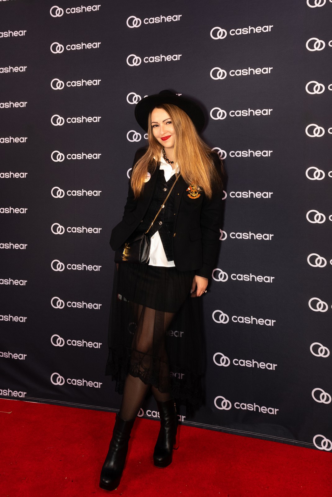 A woman in a black hat, blazer and sheer skirt poses on a red carpet before a Cashear backdrop