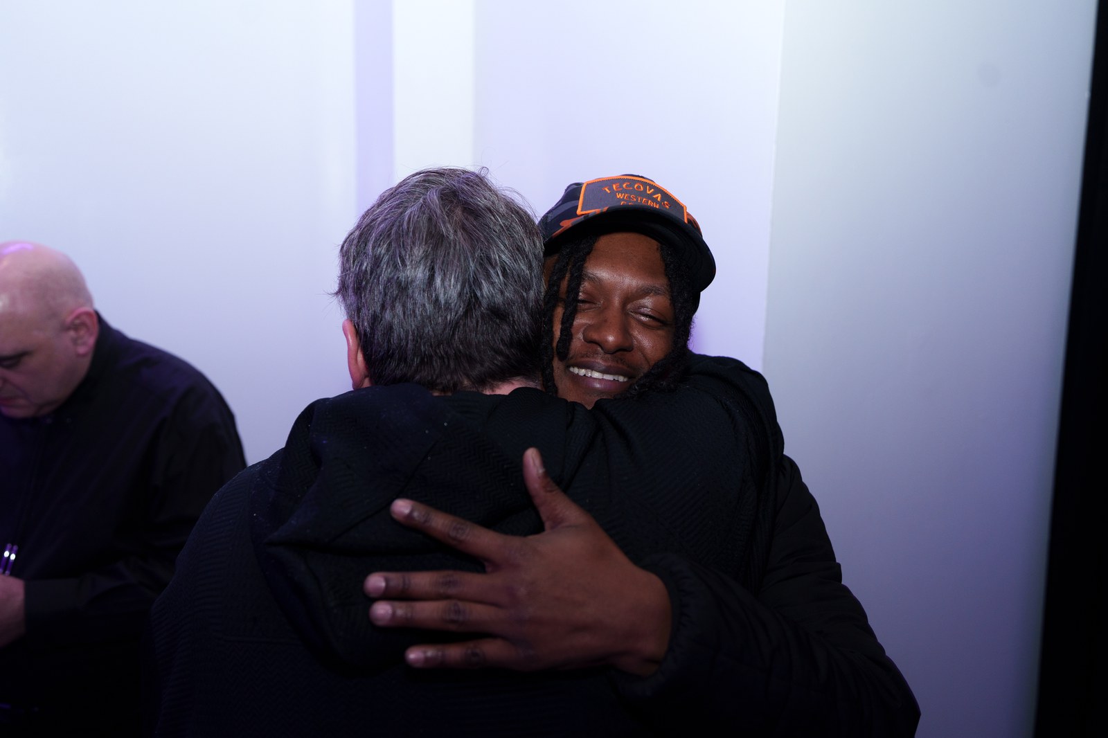 Two men in a close embrace at the launch, one in an orange cap, a pale wall behind them