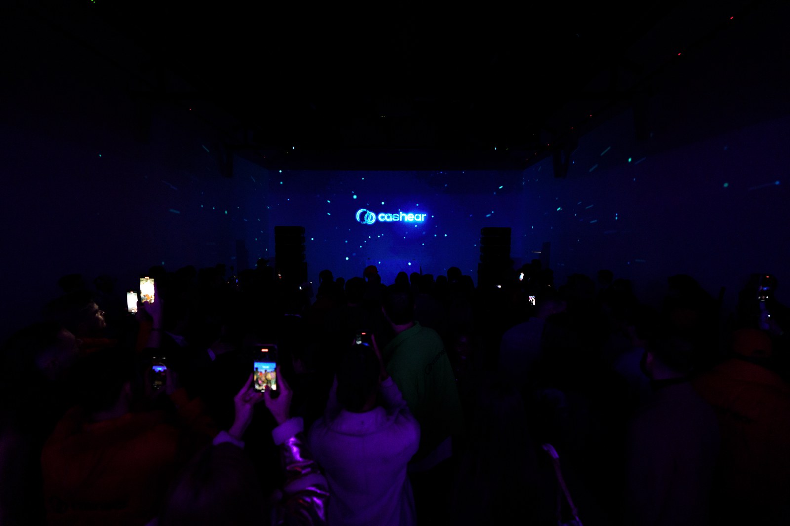 A dark crowd raises phones toward a small glowing Cashear logo on a far wall under blue light