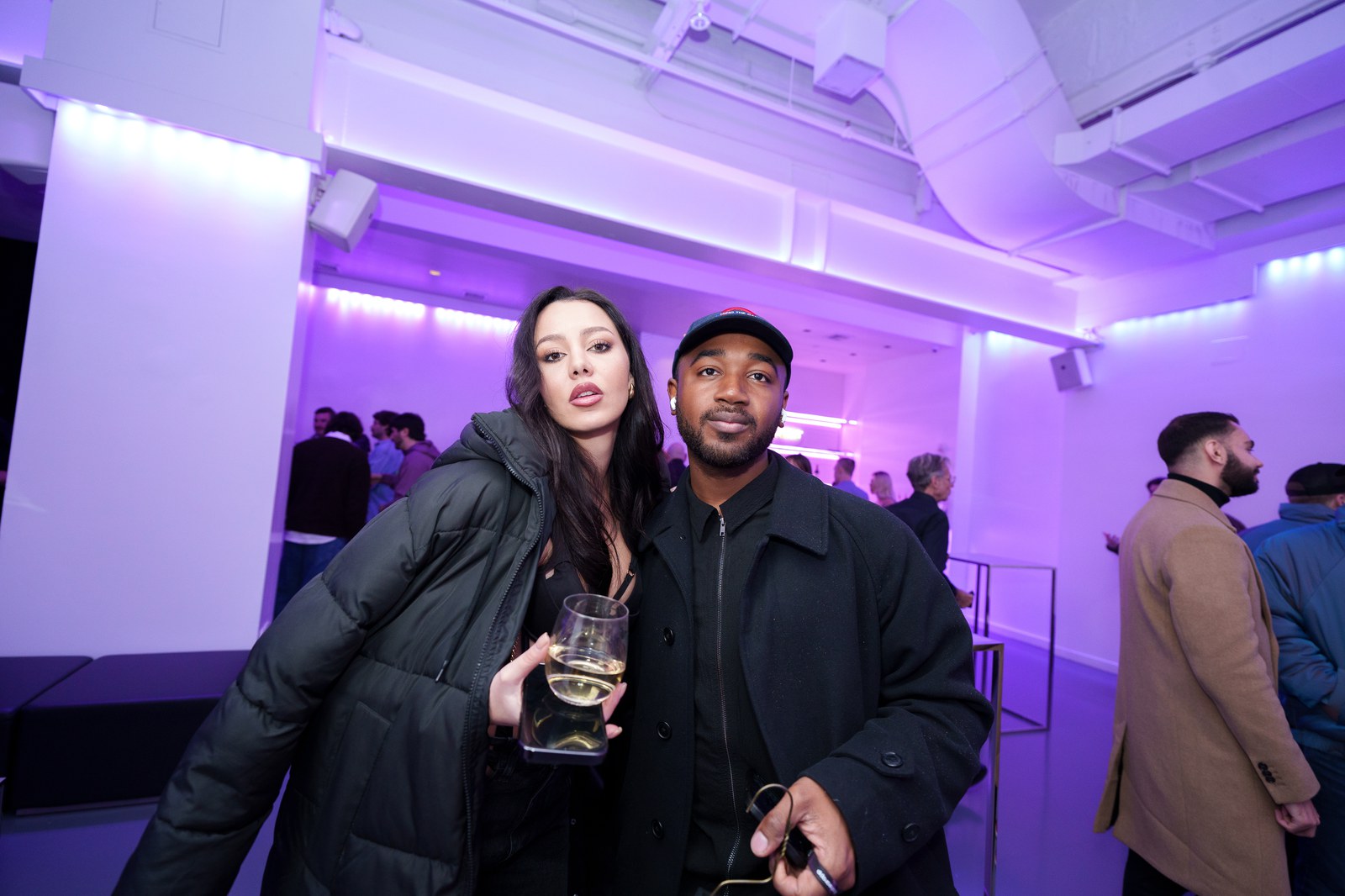 A couple posing together with drinks in a purple lit room, guests waiting behind a barrier