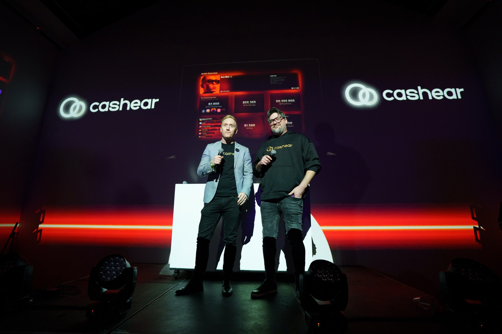Two men with microphones stand on a dark stage between projected Cashear logos and a red laser line