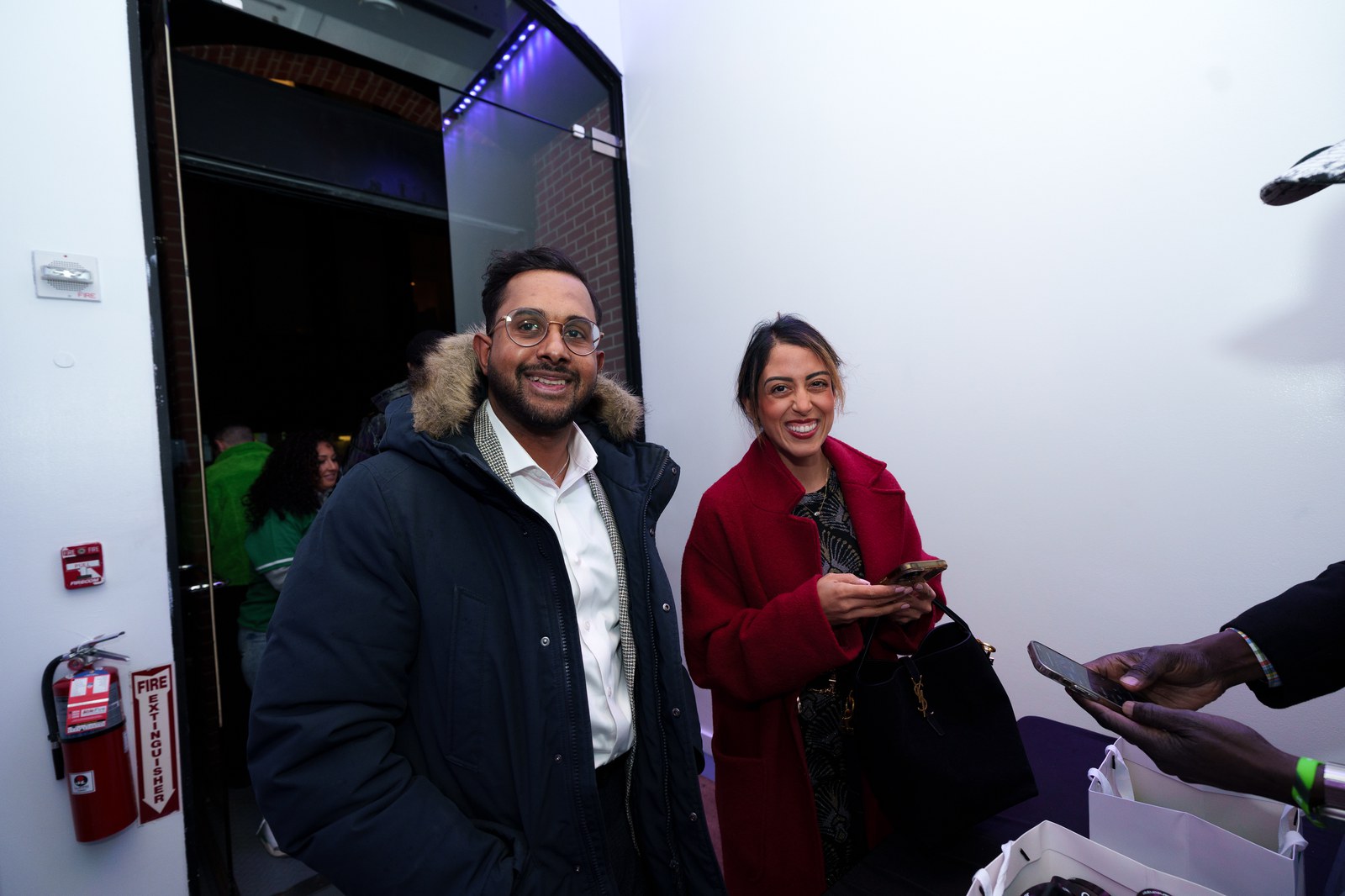 Two guests in winter coats laughing beside a doorway with green light behind them