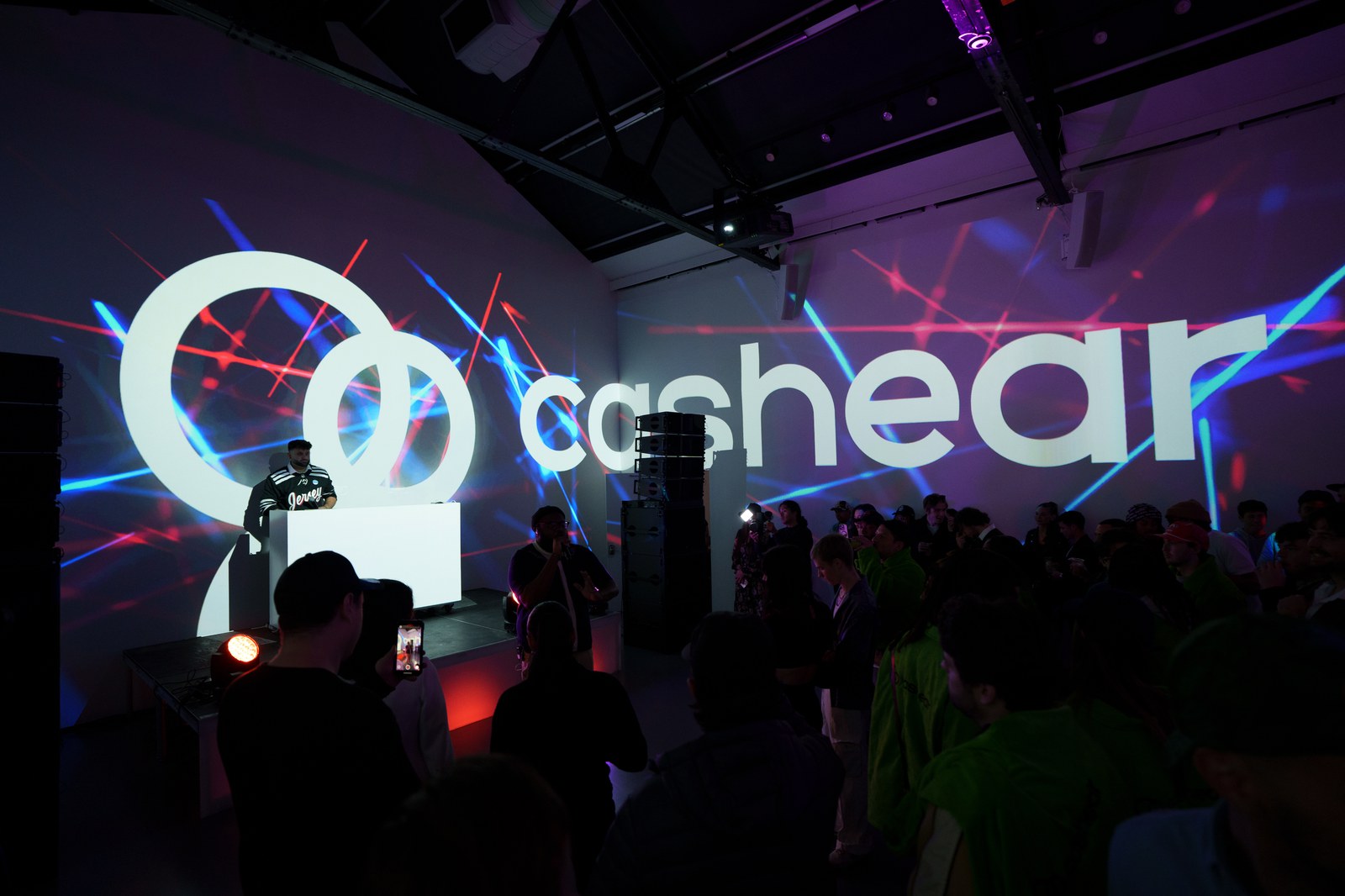 Crowd facing a DJ booth below a huge white Cashear logo crossed by red and blue laser beams