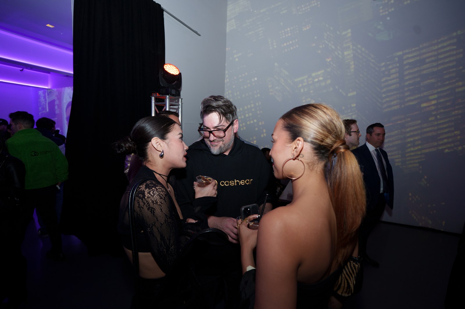 A man in a black Cashear hoodie chats with two women in front of a projected city skyline