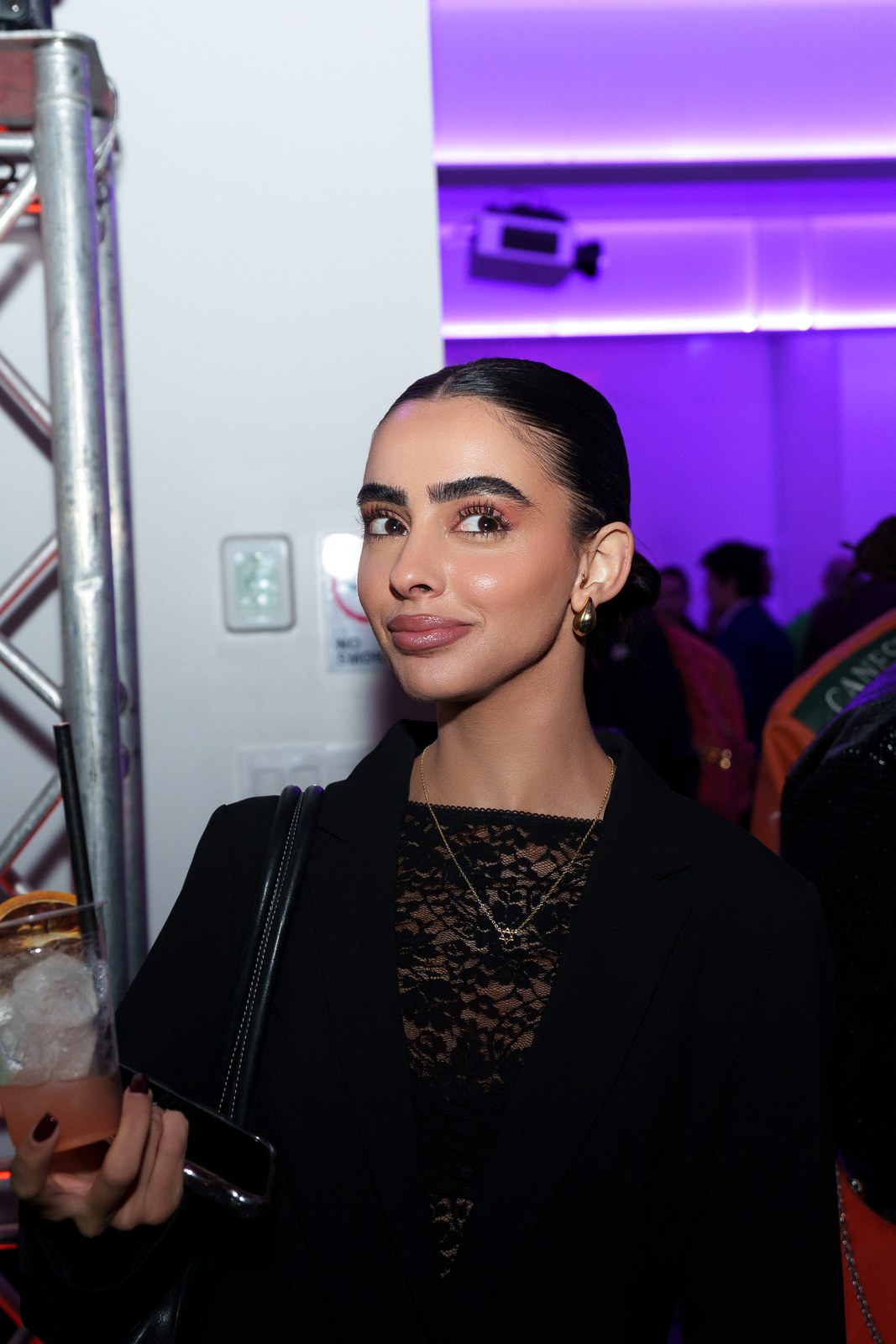 A woman in a black lace top holds a cocktail beside a metal truss in a purple lit room