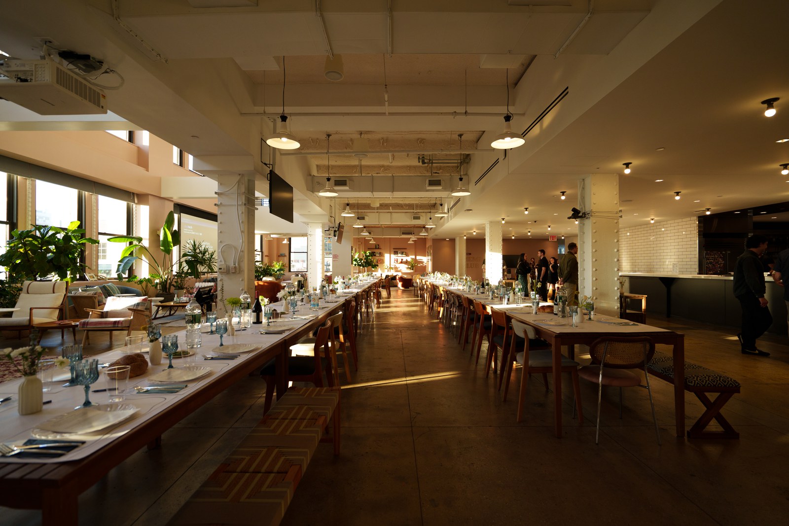 Long dining tables set with blue glassware in a sunlit loft with white columns and hanging pendant lamps