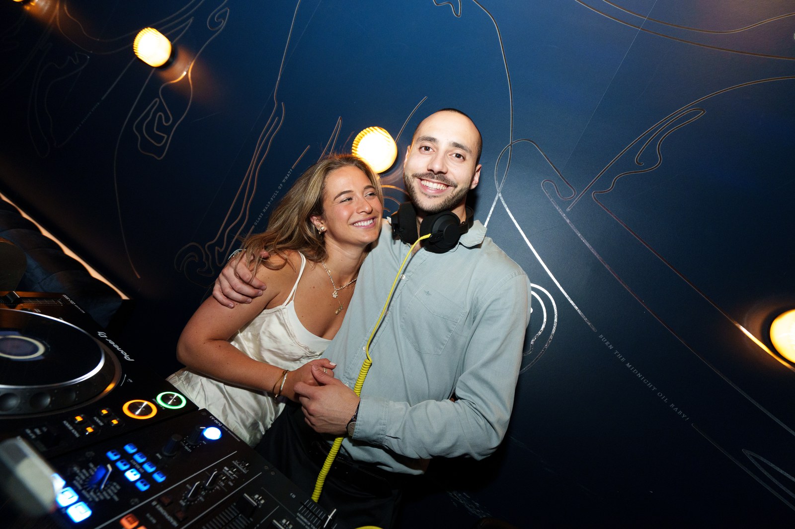 A smiling woman in white and a man wearing headphones embrace behind DJ decks under warm spotlights
