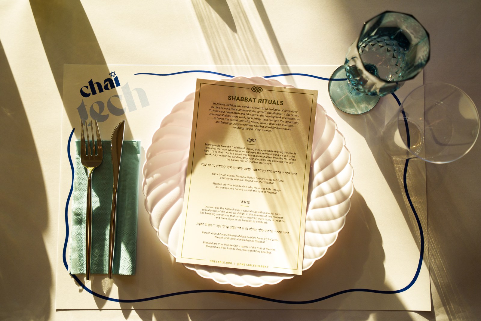 Place setting with a Chaitech placemat, Shabbat rituals card on a scalloped plate and blue glass in sunlight