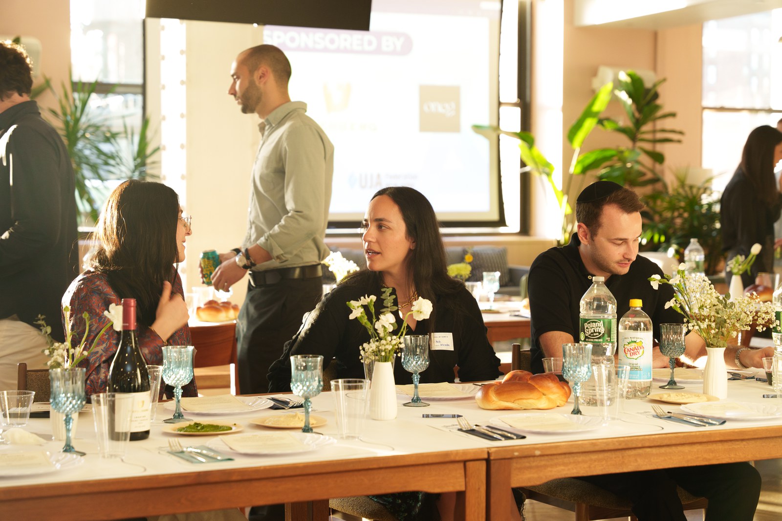 Guests seated at a long table with challah, wine and blue glasses while a man stands beside a projector screen