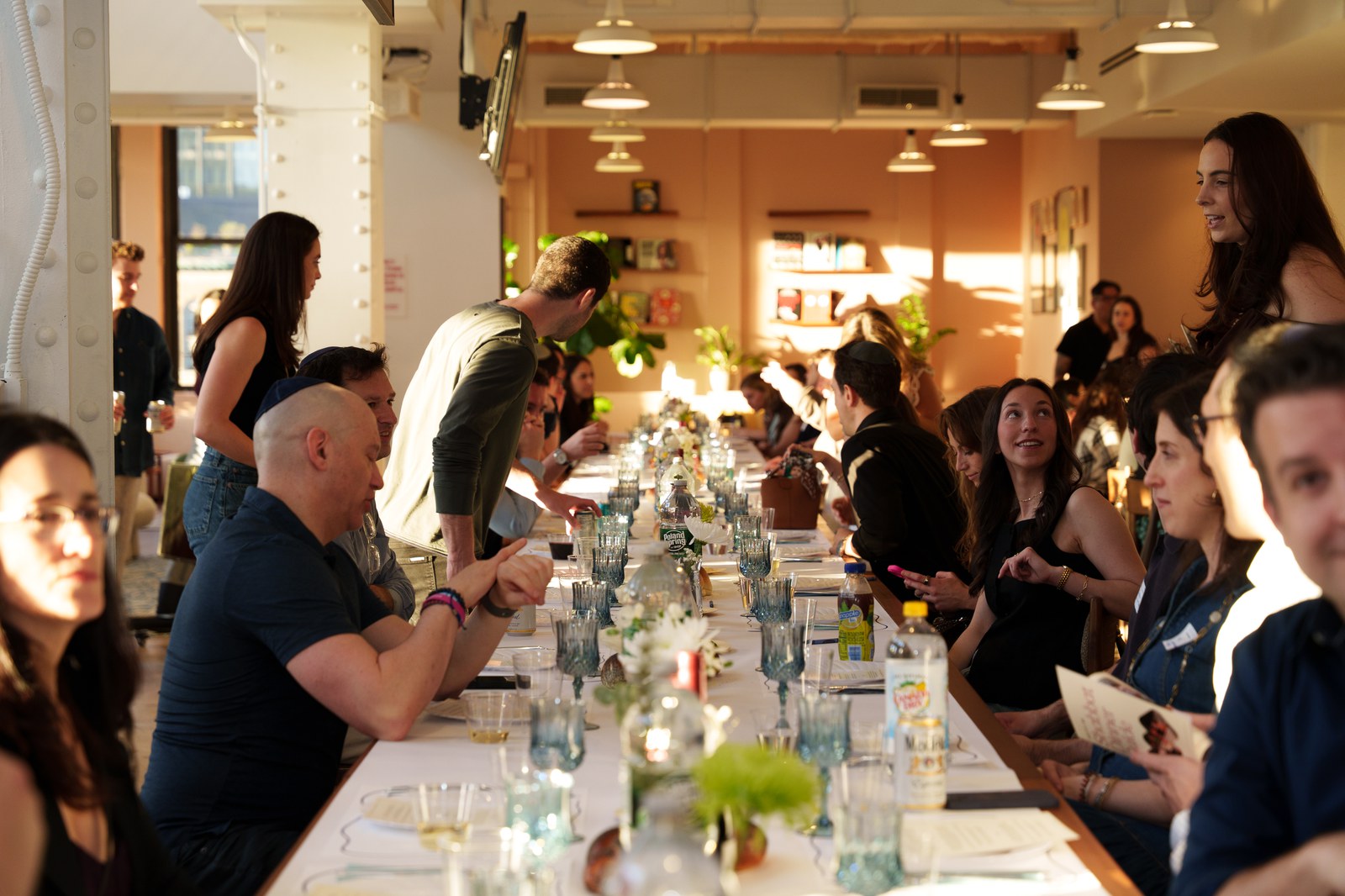 Crowded dinner along a long table in a sunlit loft, guests chatting over blue glasses and small flower arrangements