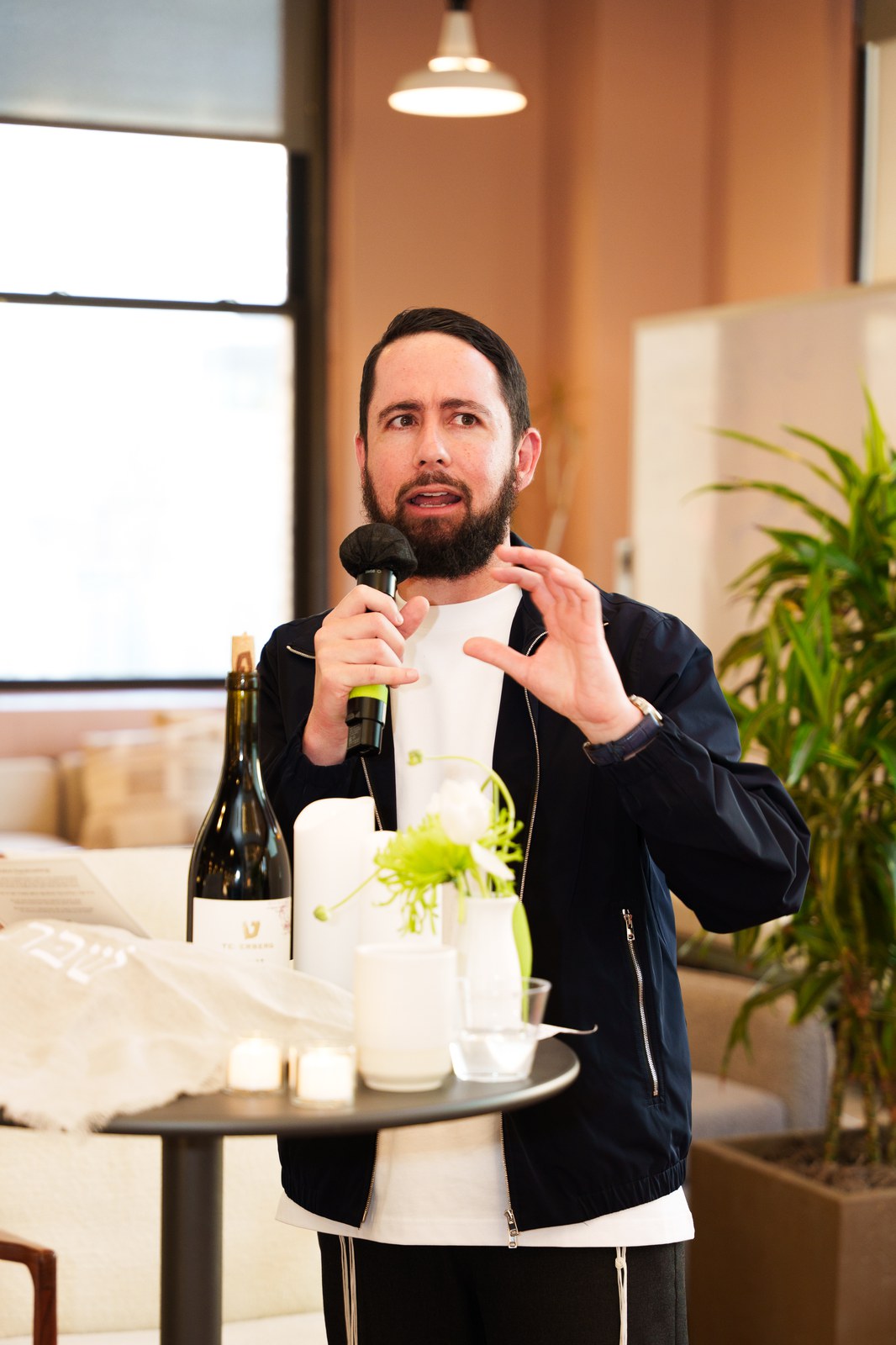 Bearded man in a navy jacket speaks into a microphone beside a small table with candles, wine and greenery