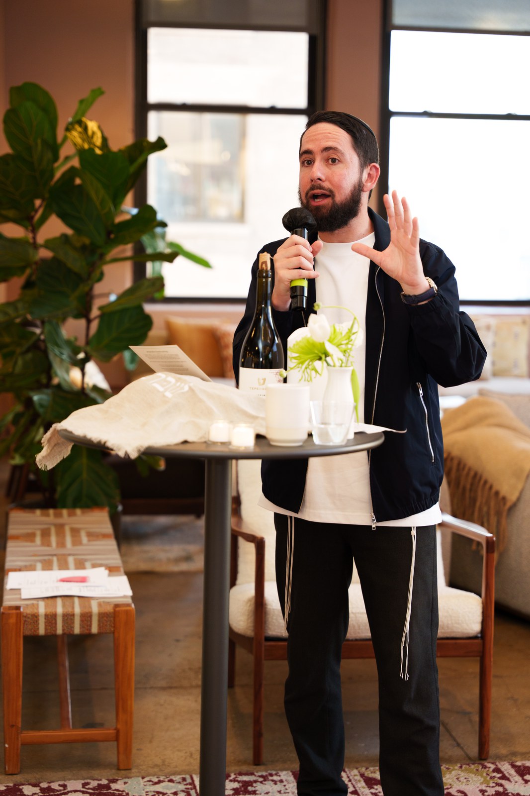 Speaker in a navy jacket raises one hand with a microphone at a round table holding wine and white candles