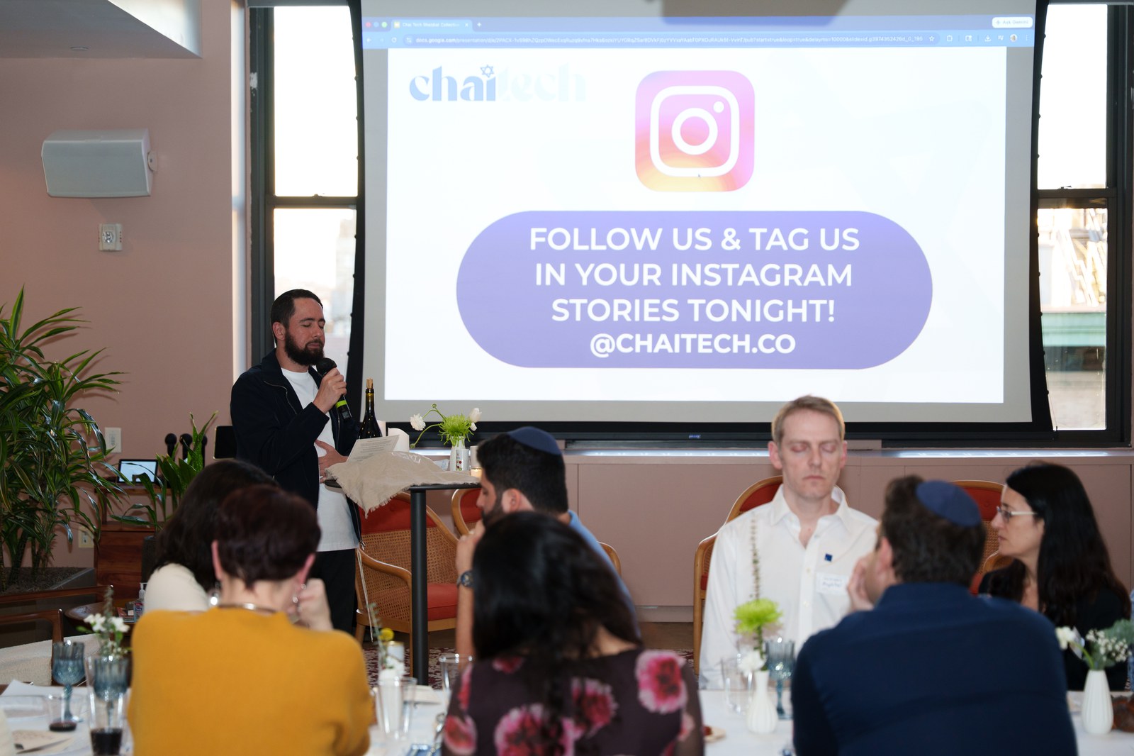 Speaker with a microphone beside a Chaitech screen asking guests to tag their Instagram stories, seated audience in front