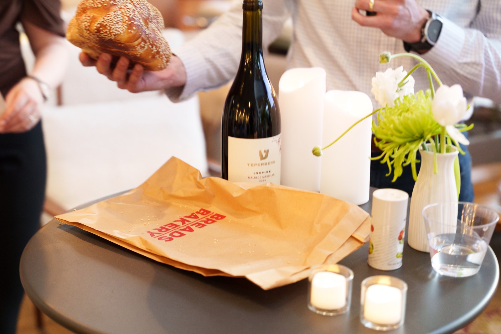 Hands hold a challah above a round table with a Breads Bakery paper bag, wine bottle and candles