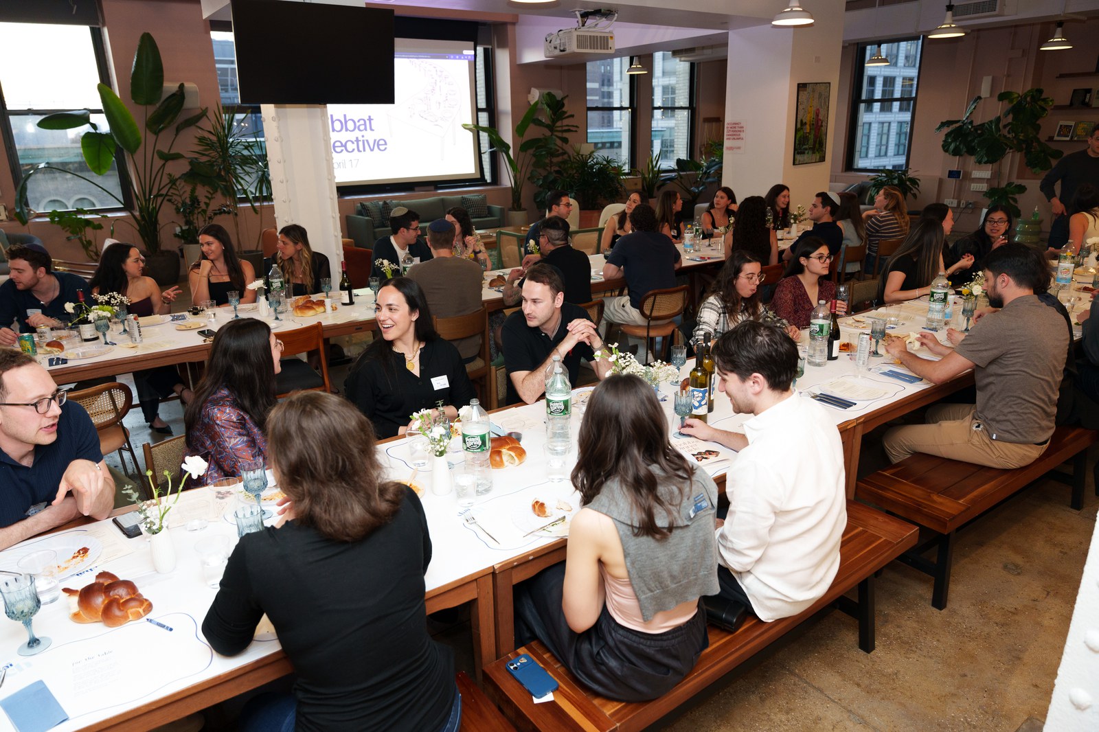 Guests dine along long tables with challah and flowers in a sunlit loft with a screen at the far wall