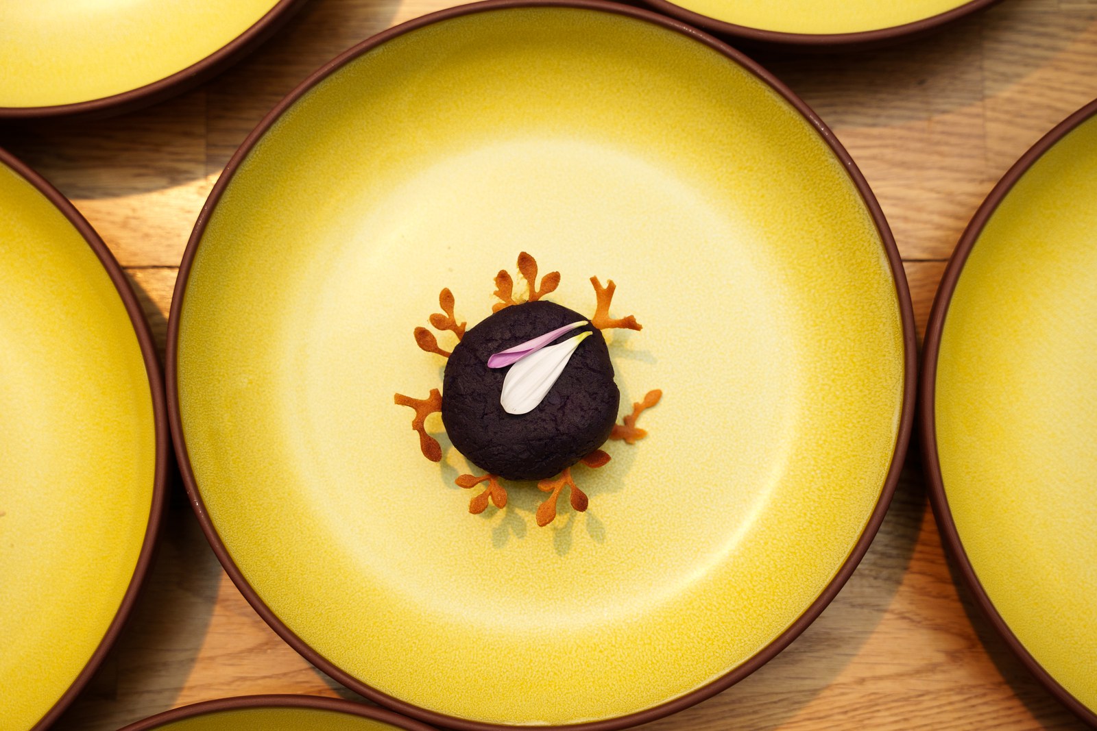 A dark round bite topped with a purple petal sits centred on a yellow plate among matching plates
