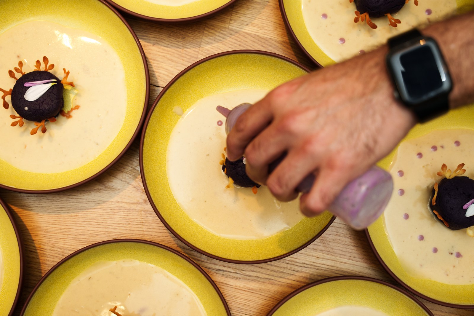 Overhead view of a hand piping sauce beside dark purple spheres on yellow rimmed plates on a wooden table