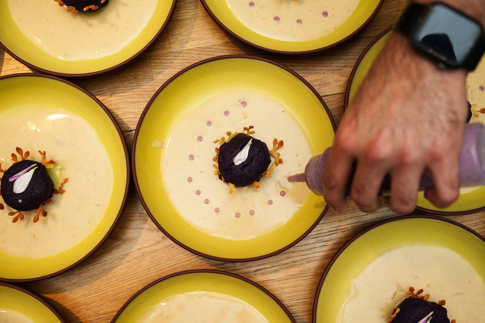 Overhead view of yellow rimmed plates with dark purple domes as a hand squeezes sauce dots around one