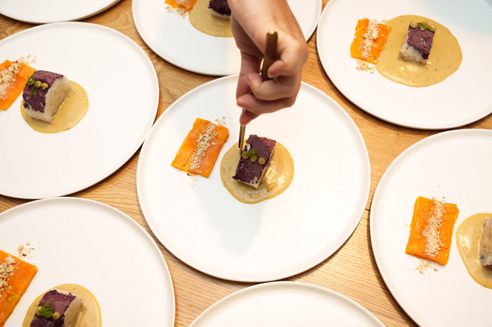 Hand plating a purple and cream dessert on white plates with an orange tuile on a wooden table