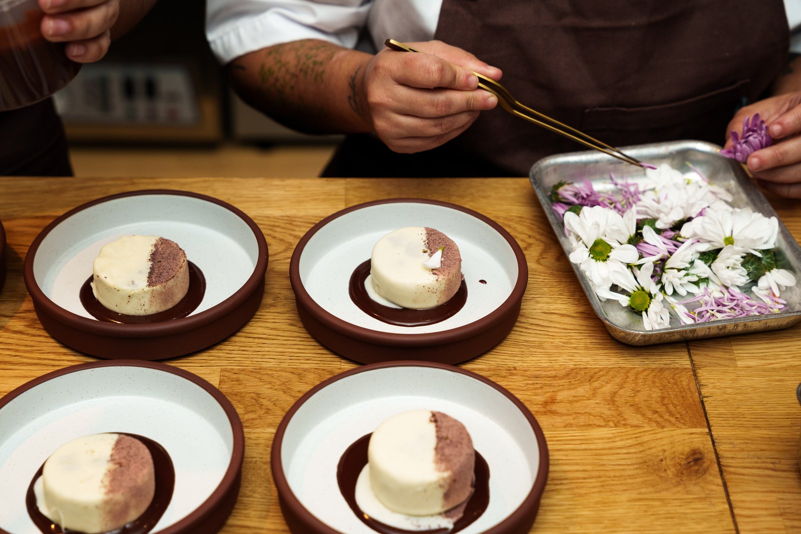 A chef uses tweezers to place edible flowers on rounds of cream dessert plated in terracotta bowls