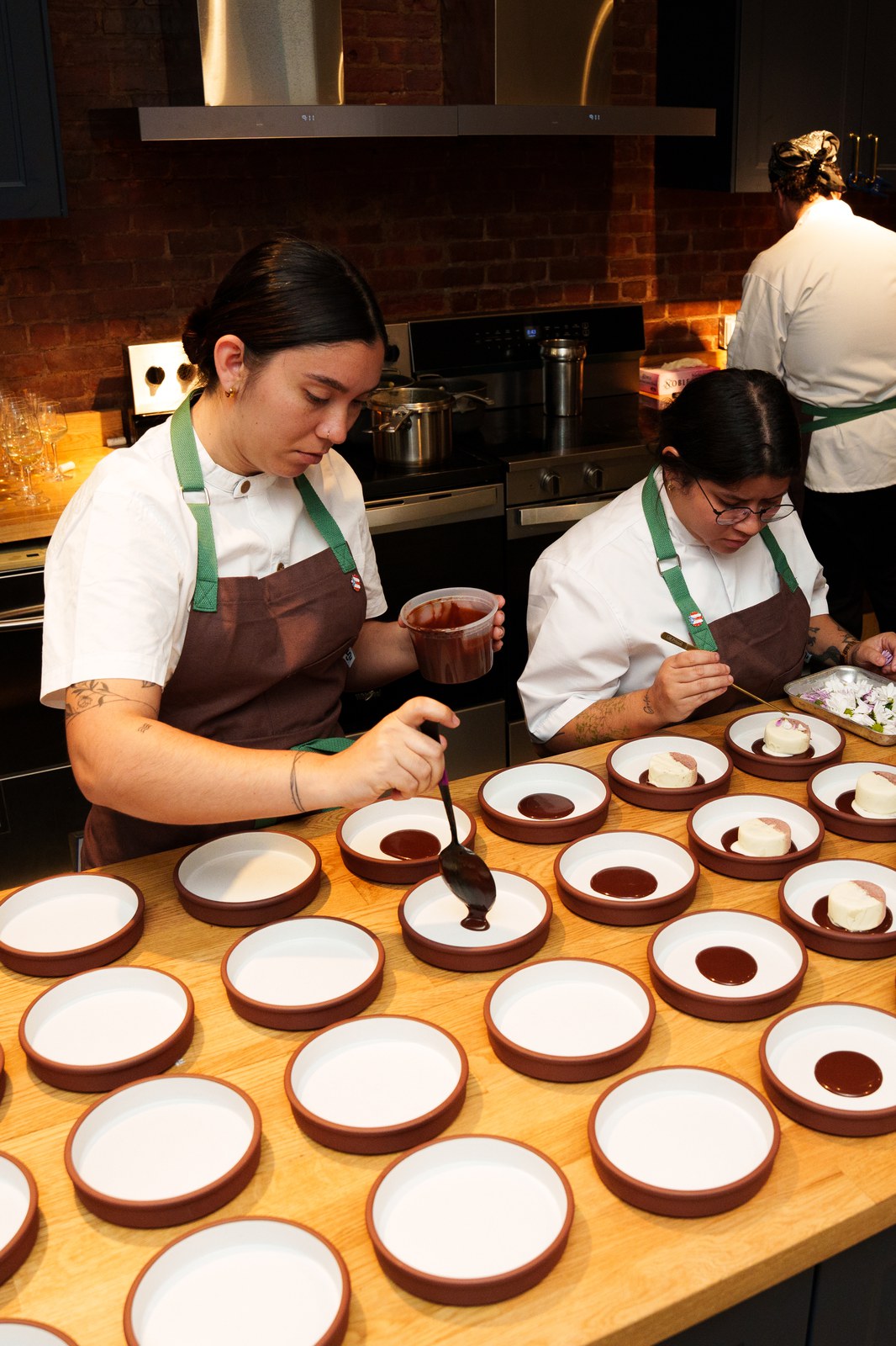 Two cooks in brown aprons spoon dark sauce into rows of white and terracotta plates on a wooden counter