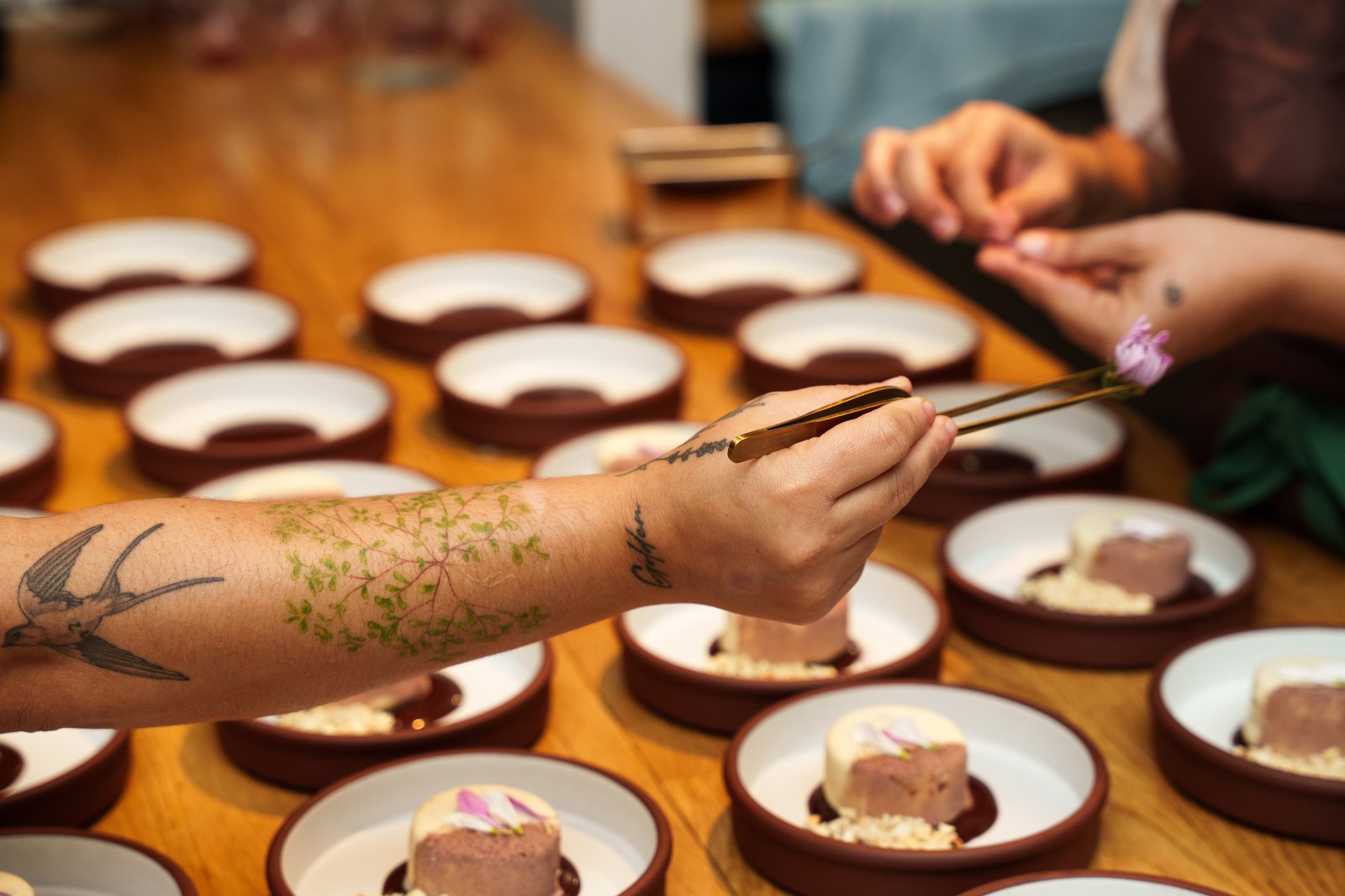 Tattooed arm placing a petal with tweezers on rows of small desserts in terracotta dishes
