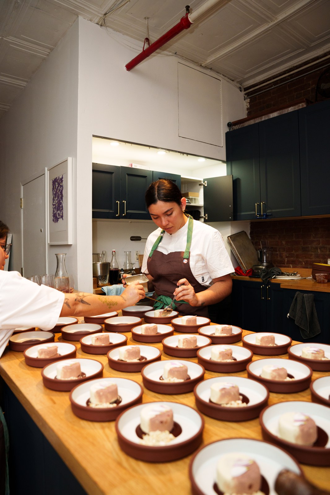 A chef in a white jacket and brown apron plates dozens of small desserts in a brick walled kitchen