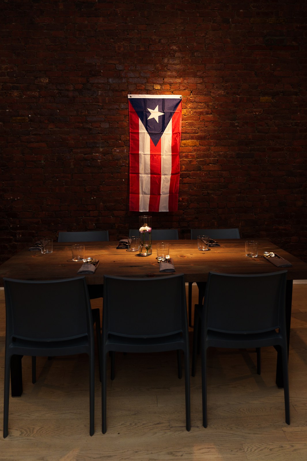 A wooden dining table with black chairs set beneath a Puerto Rican flag on a lit brick wall