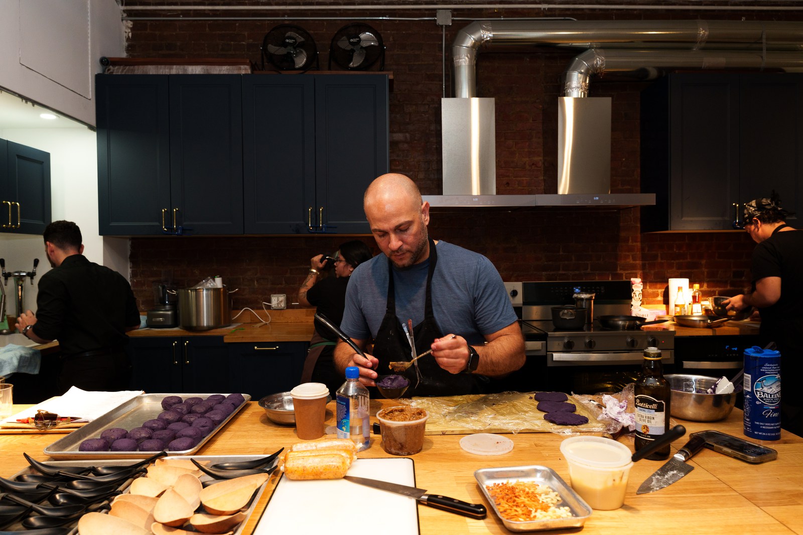 Bald chef in a blue tee and black apron preparing purple dough balls in a brick walled kitchen