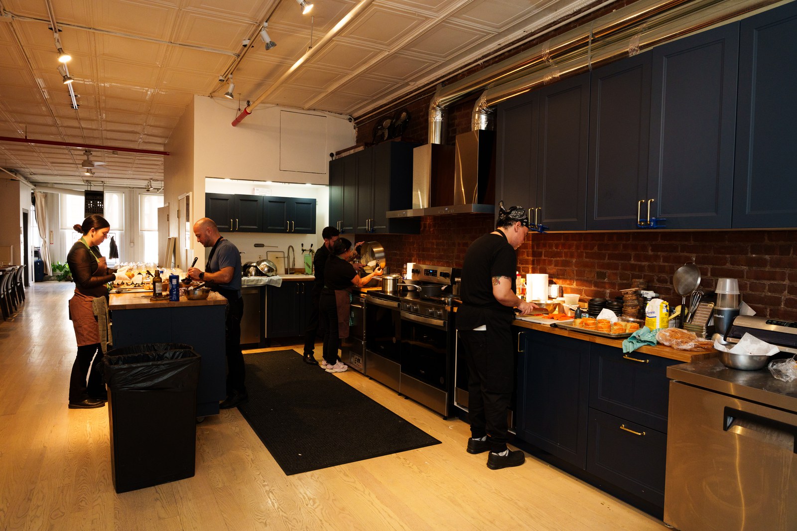 Cooks work at a navy blue kitchen with brass fittings and a brick backsplash in a loft space