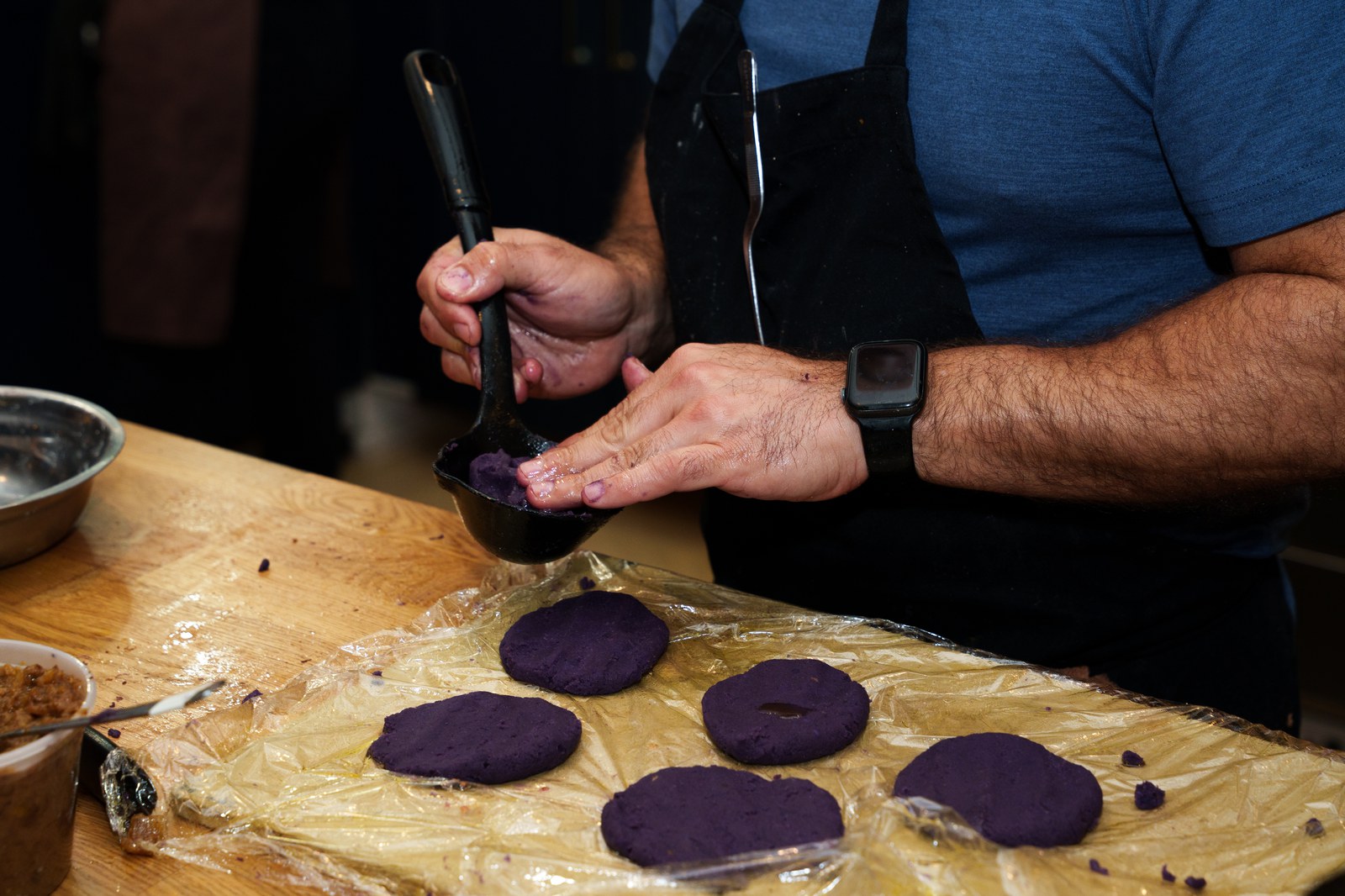 A cook in a black apron presses purple dough into flat discs on plastic wrap on a wooden counter