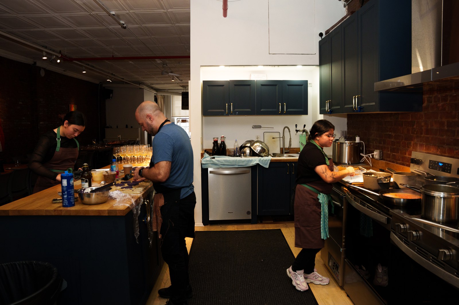 Three cooks in aprons working at counters in a navy cabinet kitchen with a brick wall and steel range