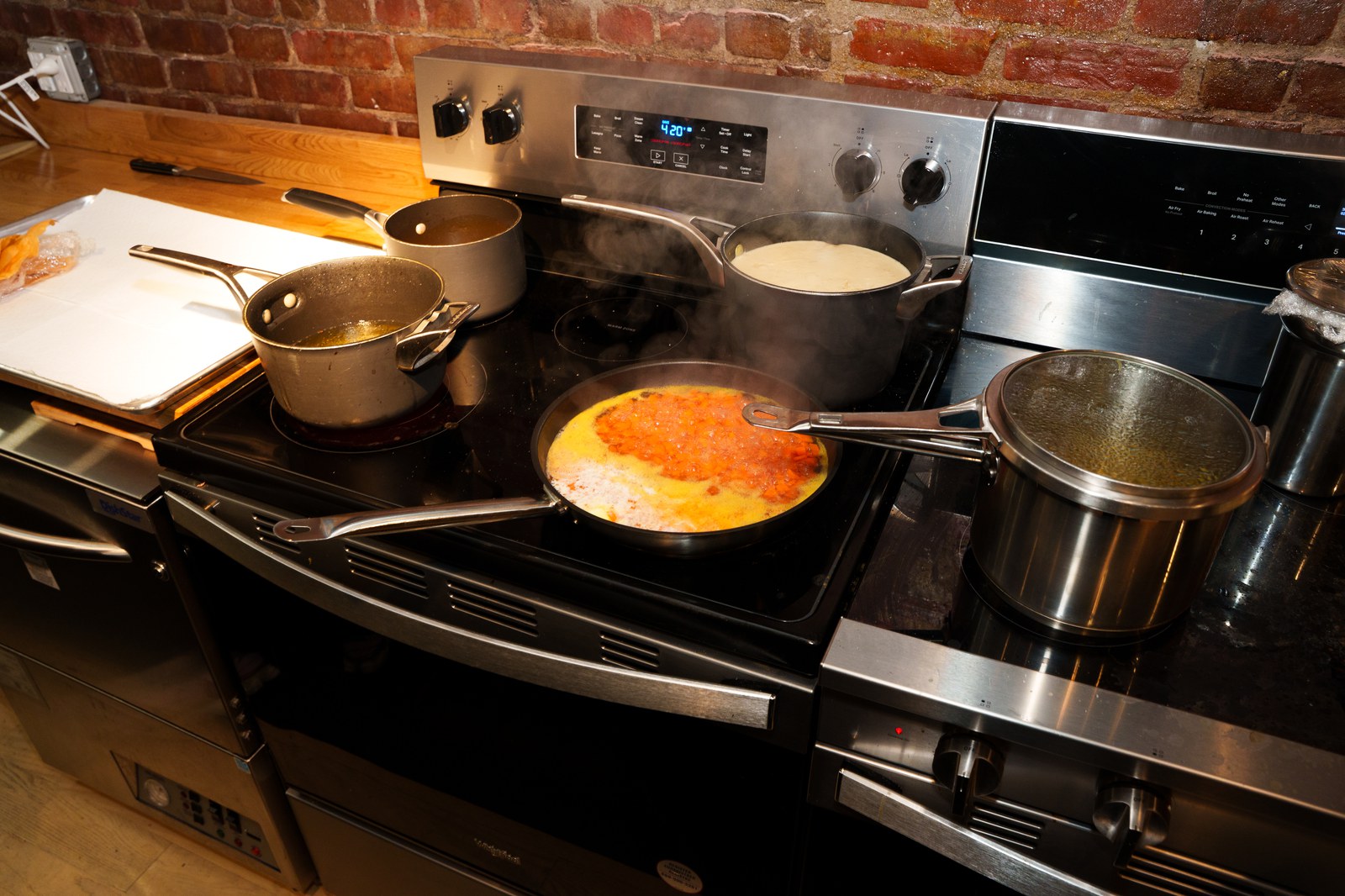Steaming pots and a pan of orange sauce crowd a stainless stove in a brick walled kitchen