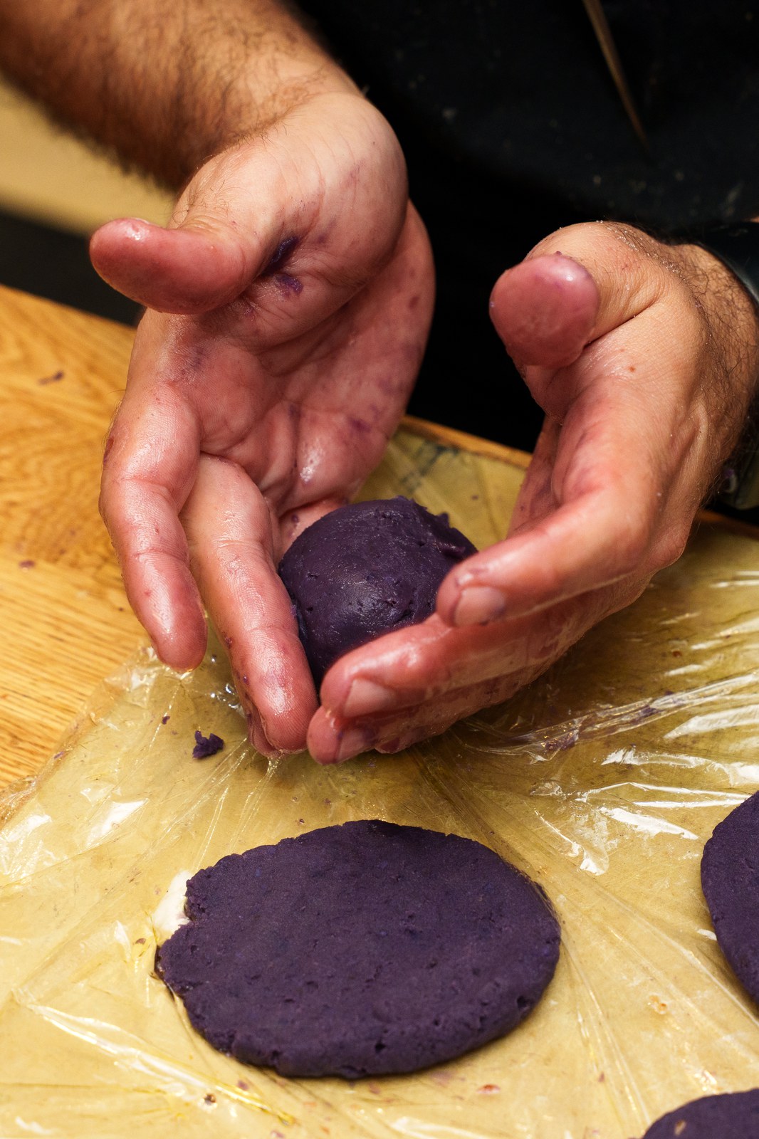 Purple stained hands shape a ball of dark purple dough over a plastic wrapped wooden board