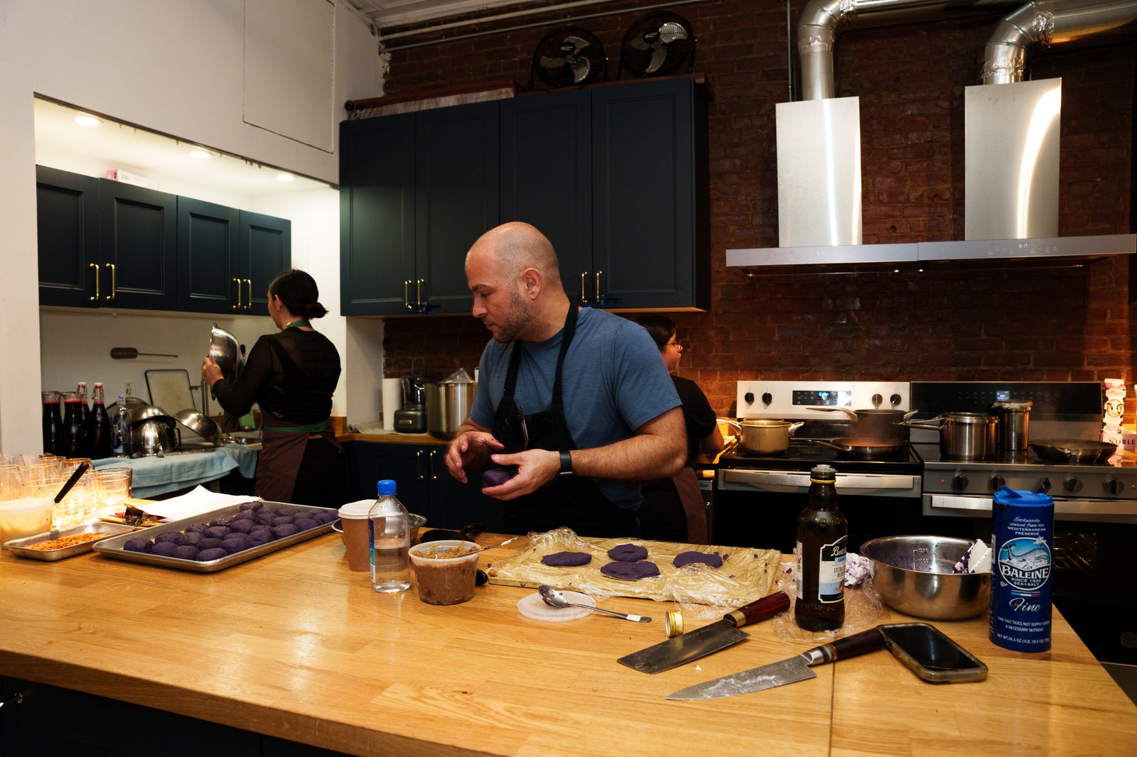 A cook in a blue shirt and black apron works purple dough at a wooden kitchen island