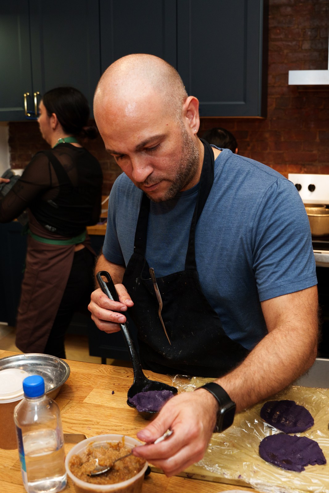 Chef in a black apron portioning purple dough on plastic wrap beside a tub of filling