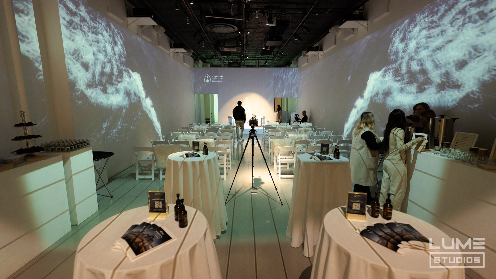 White room with cocktail tables, folding chairs, a camera on a tripod and swirling water projected on the walls
